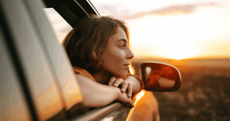 A young woman with curly hair leans out of a car window, resting her arms on the door frame while looking toward a bright, golden sunset.