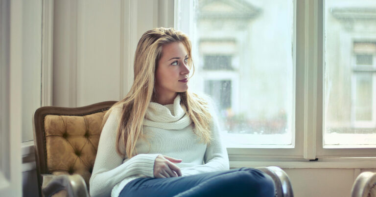 Blonde woman in a white cowl-neck sweater and jeans sitting in an armchair, looking out a large window.