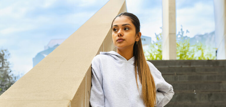 Young woman in a gray hoodie standing next to a concrete structure outdoors.